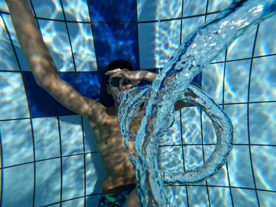 A person lying at the bottom of a pool holding their nose, blowing out air bubbles that look like a vortex when viewed from above.