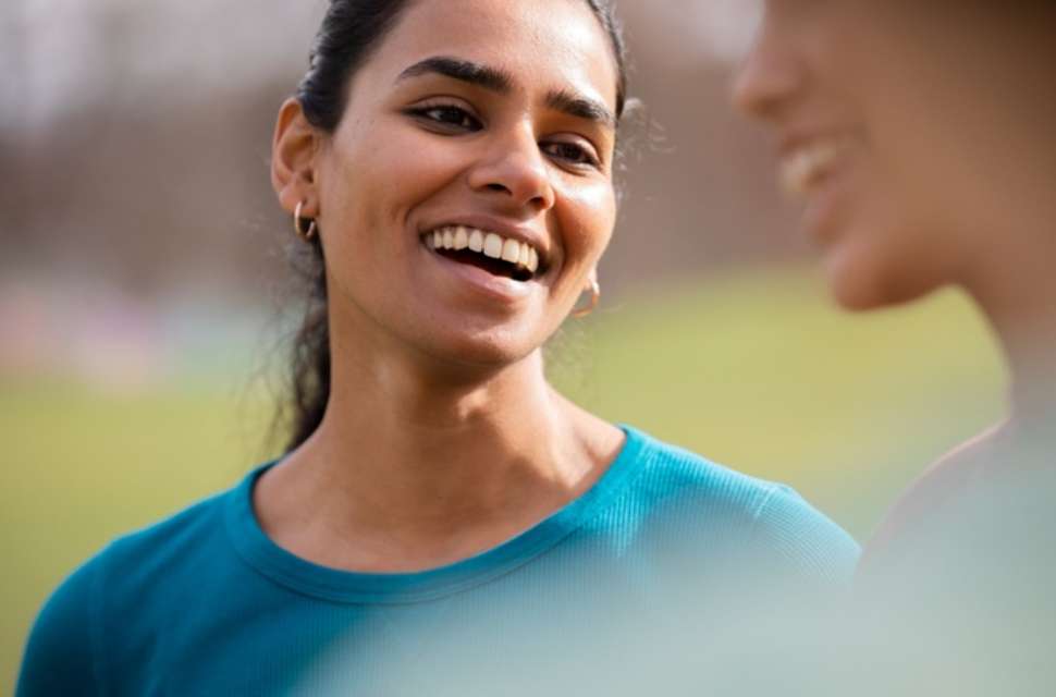 A young woman smiling warmly while talking to someone outdoors. She appears relaxed and engaged, with a soft natural background behind her.