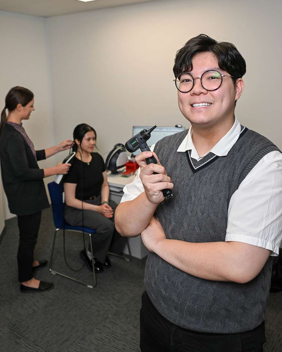 Flinders Audiology student stood smiling with tool with patient in the background