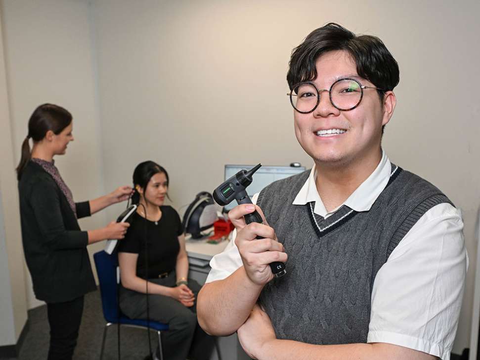 Flinders Audiology student stood smiling with tool with patient in the background
