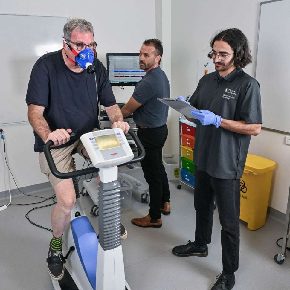 Man on exercise bike doing testing with a mask on with flinders exercise physiology student and teacher next to him