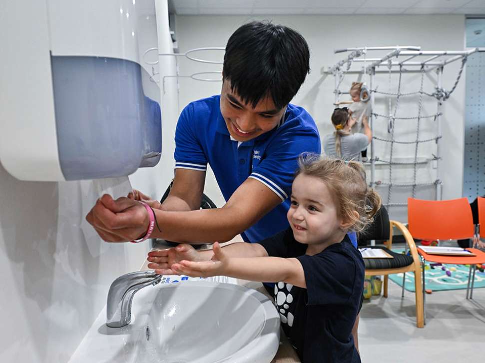 A speech pathology student and a young child standing at a sink, washing their hands together.