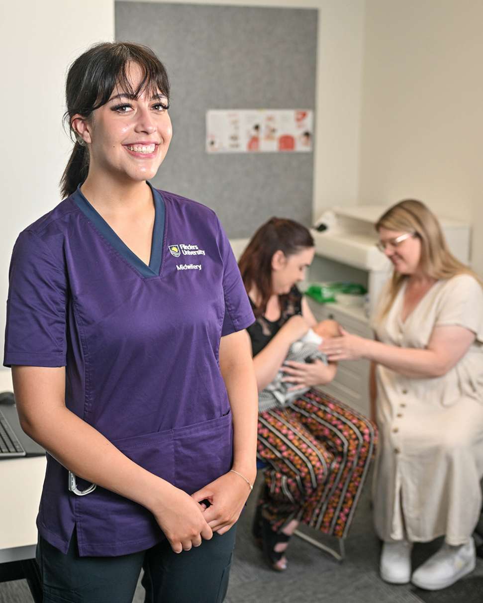 A Flinders Midwifery Student in a purple shirt stands beside a woman in a clinic, providing care and support.