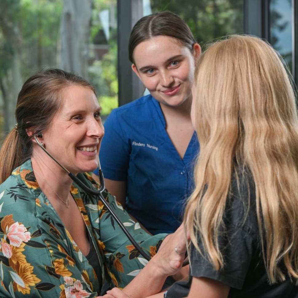 Nurse smiling and checking child's heart with Flinders Nursing student smiling and sat behind