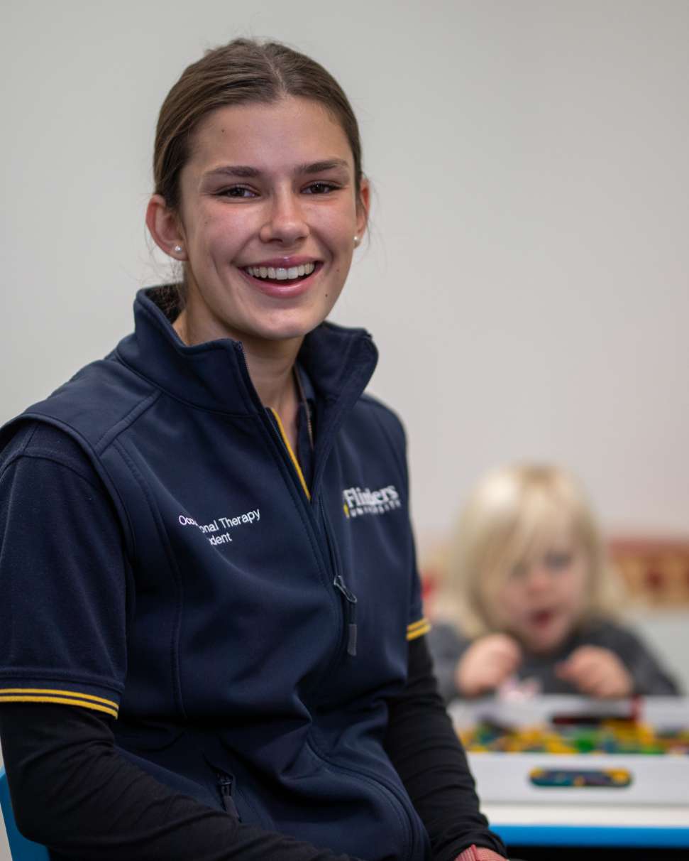 Flinders Occupational Therapist smiling at camera with child playing with toys behind