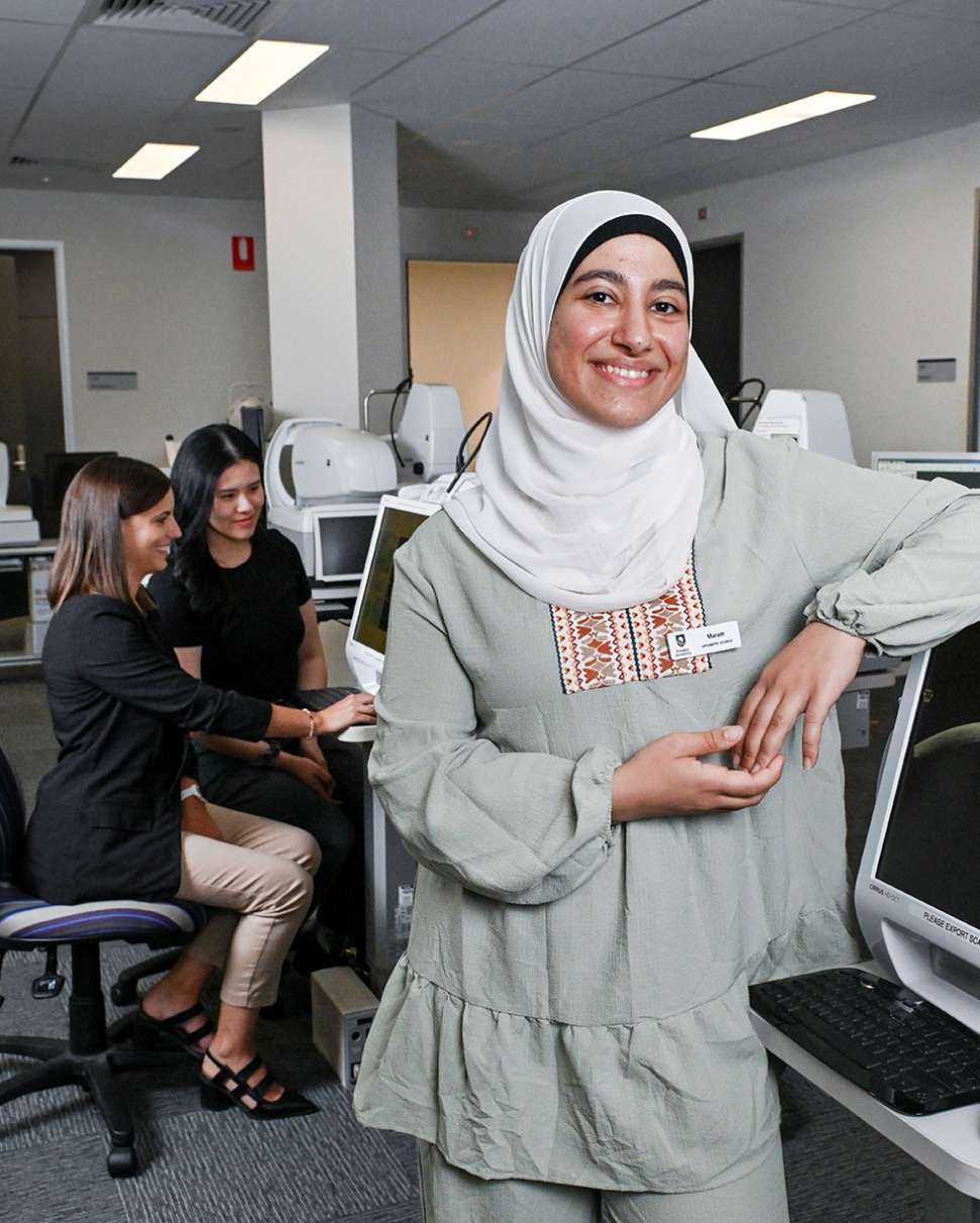 Flinders Optometry Student smiling at camera with arm lent on computer screen with patient and clinician in background