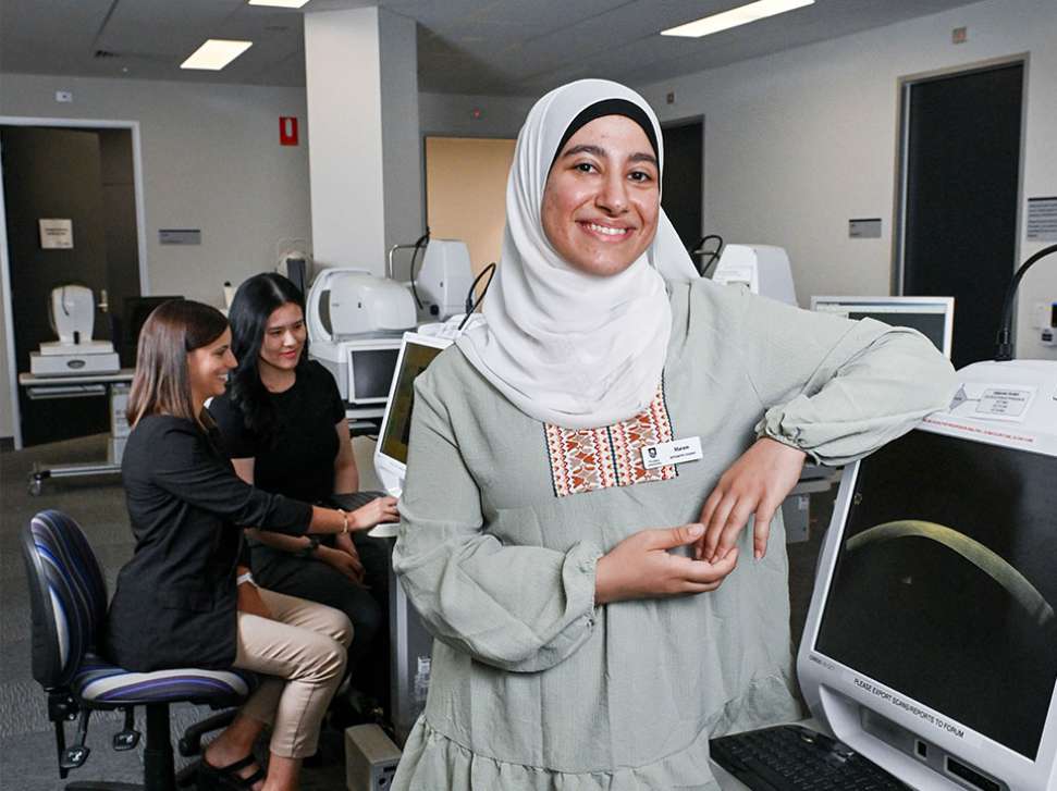 Flinders Optometry Student smiling at camera with arm lent on computer screen with patient and clinician in background