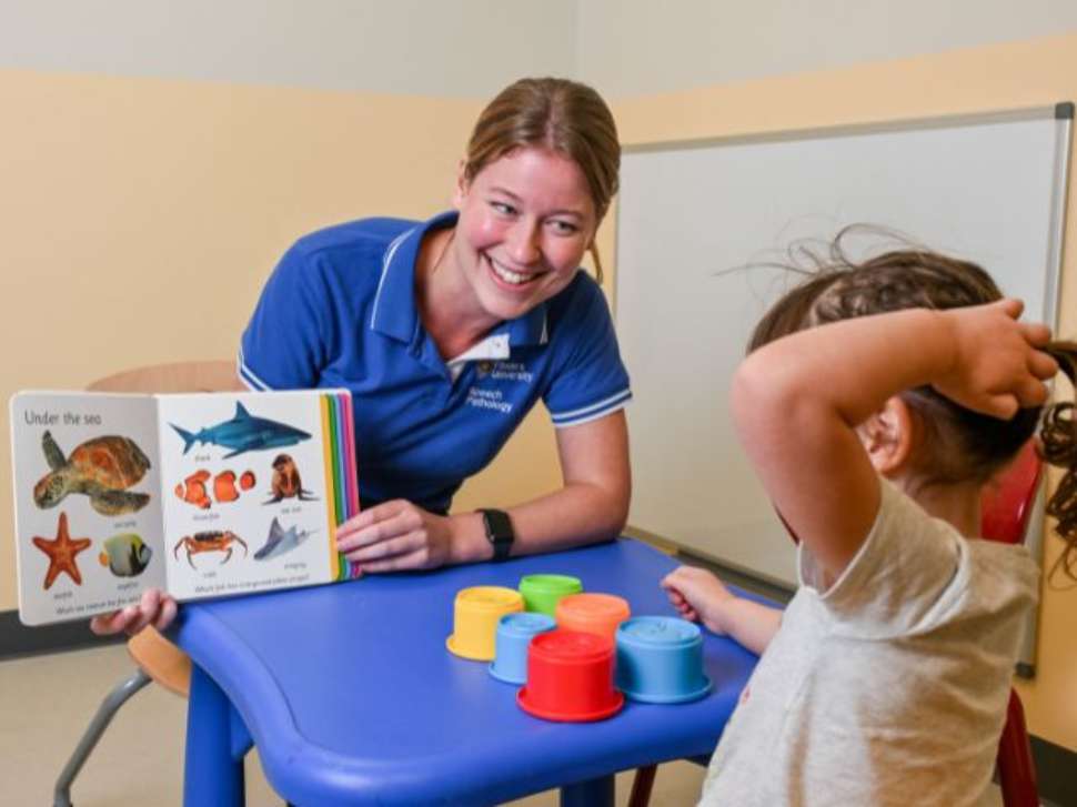 Flinders Speech Pathology Student smiling and holding up a book with pictures of animal with child looking at it