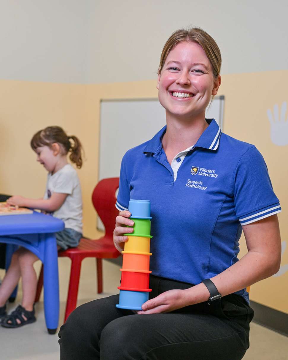 Flinders Speech Pathology Student smiling and holding colourful stacking toy with child in background