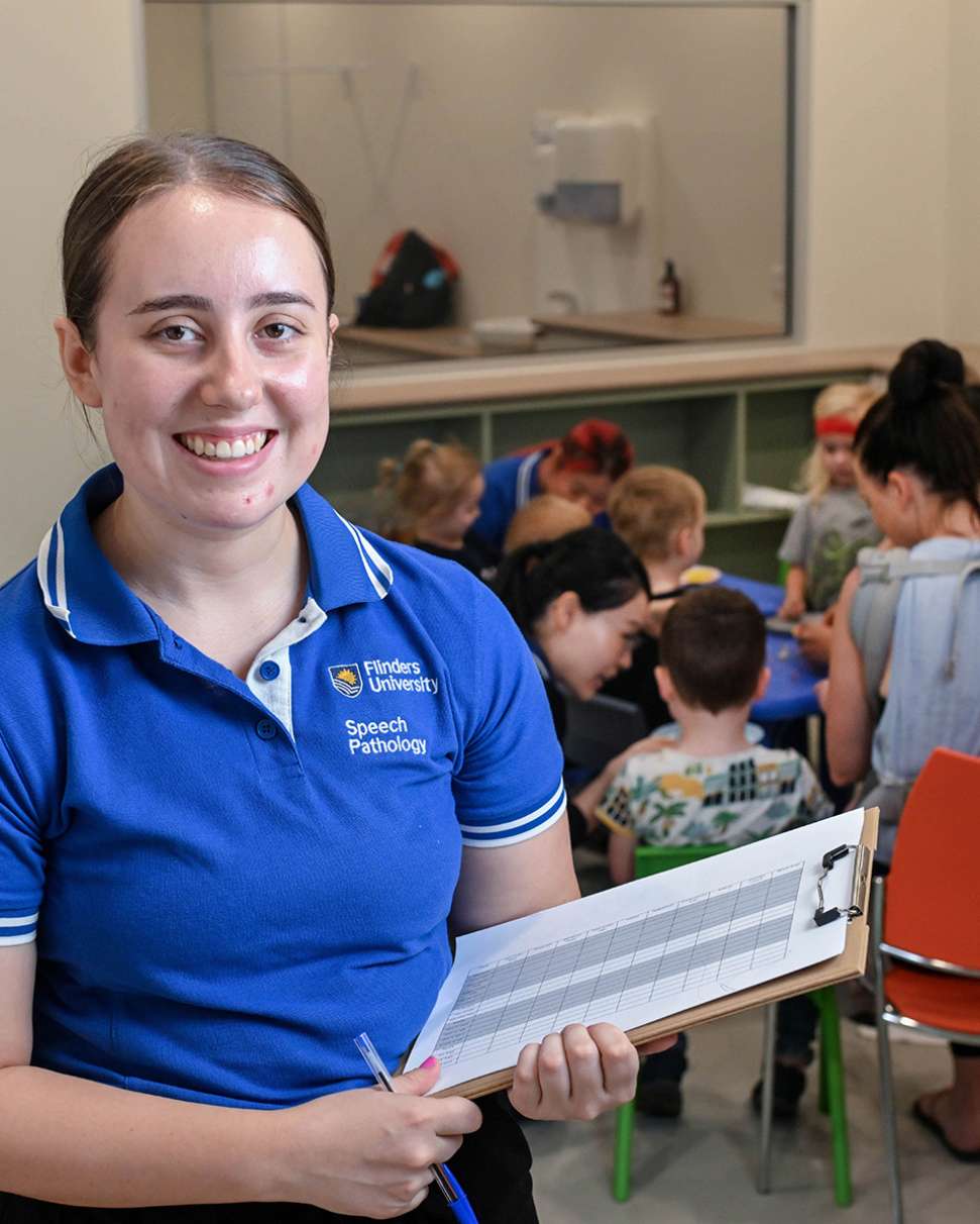 Flinders Speech Pathology Student smiling and holding clipboard with children behind