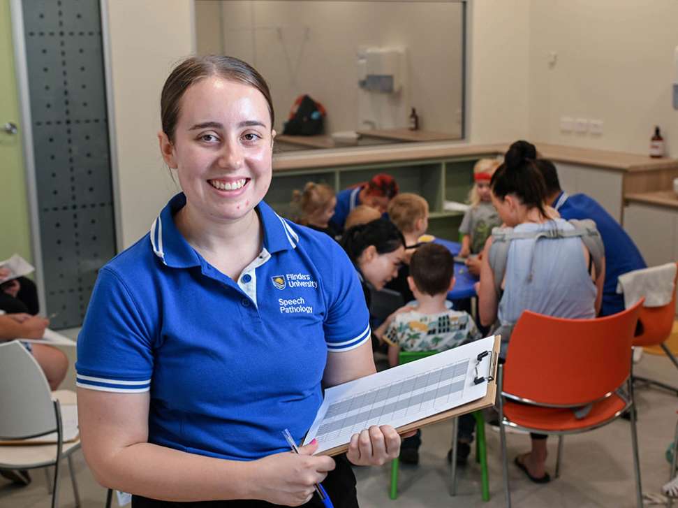Flinders Speech Pathology Student smiling and holding clipboard with children behind