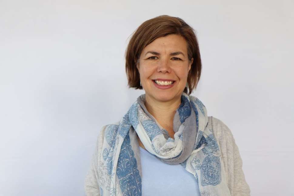 Smiling woman standing in front of a Flinders University media wall, wearing a light blue scarf and beige cardigan.